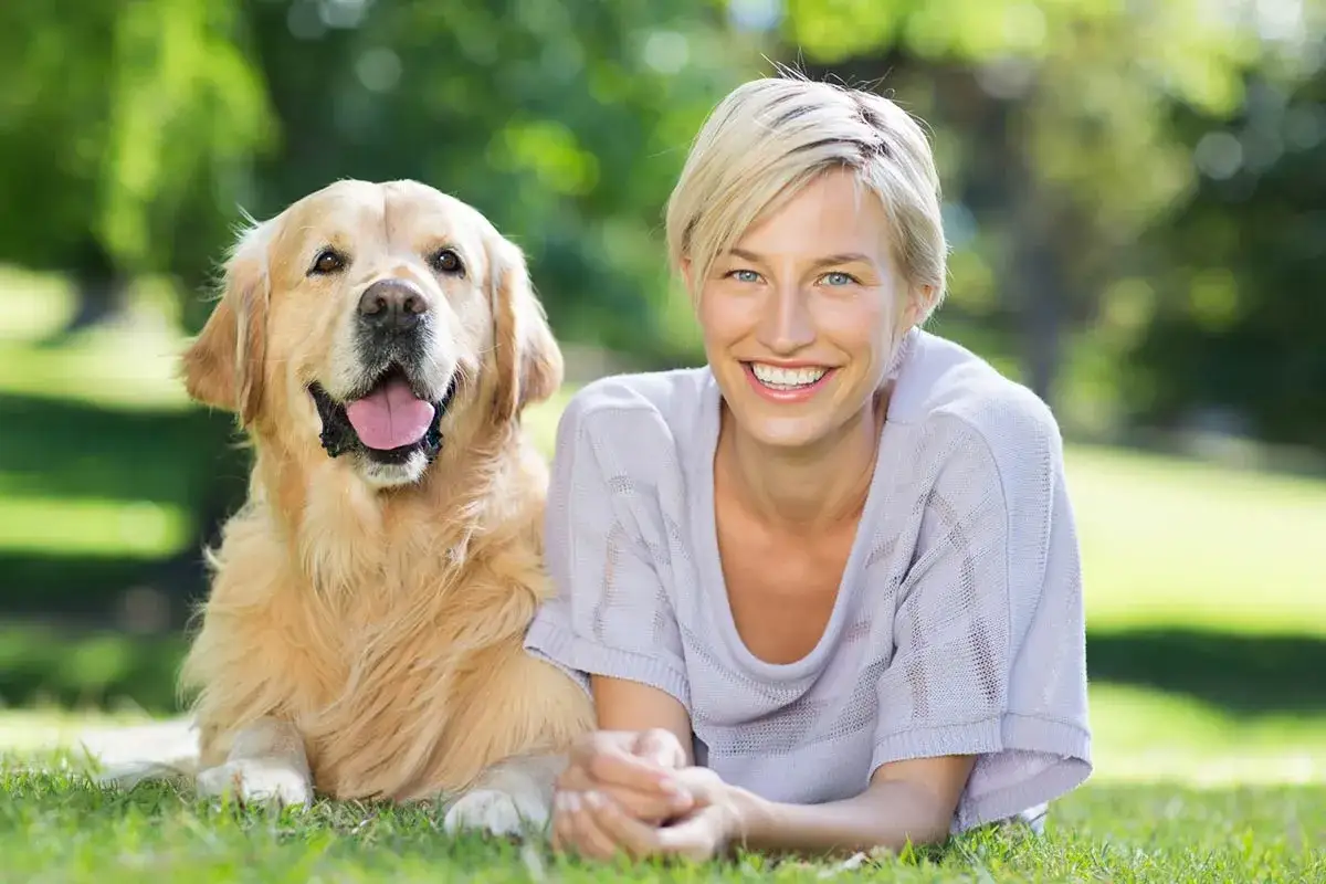 Uśmiechnięta kobieta i jej złoty retriever, jedne z najmądrzejszych ps&oacute;w, leżą na trawie w parku.