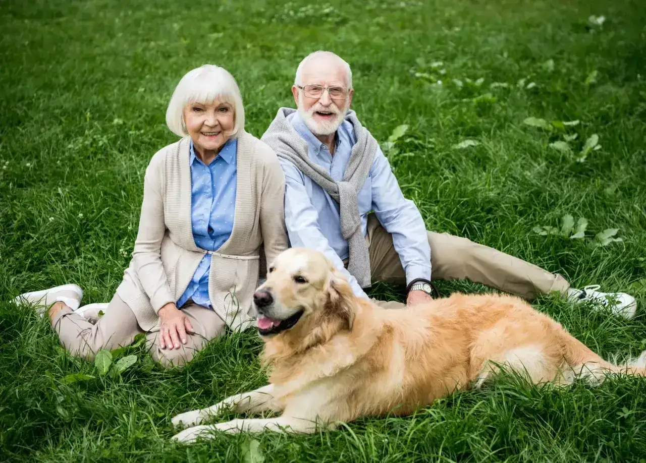 Uśmiechnięci seniorzy siedzą na trawie z psem. Złoty retriever leży obok, idealny pies dla seniora, kt&oacute;ry wnosi radość i towarzystwo.
